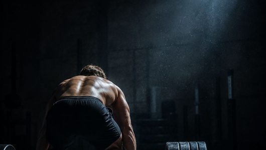 Athlete standing at a loaded barbell in a dark gym at dawn, ready to begin a hybrid training session