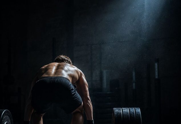 Athlete standing at a loaded barbell in a dark gym at dawn, ready to begin a hybrid training session
