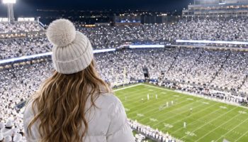 Penn State game day outfits for the White Out: a woman seen from behind in a white puffer jacket, white pom-pom beanie and light jeans above a night game where the whole crowd wears white