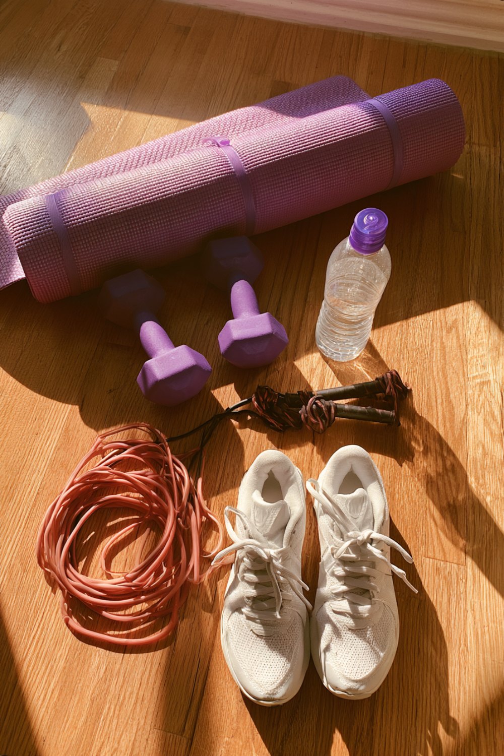 Home workout gear on a wood floor: purple yoga mat, dumbbells, jump rope, water bottle and white sneakers