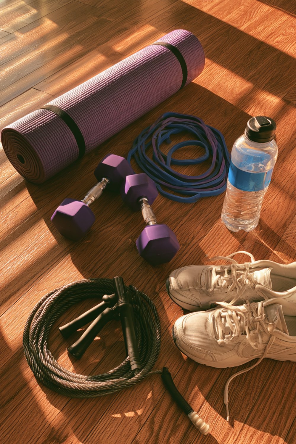 Rolled yoga mat, hex dumbbells, resistance band, jump rope and sneakers in morning sun