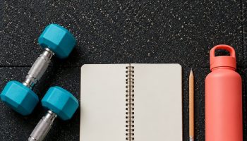 Flat lay of teal dumbbells, a blank notebook, water bottle, resistance band and four colored notes for a 4 day workout split