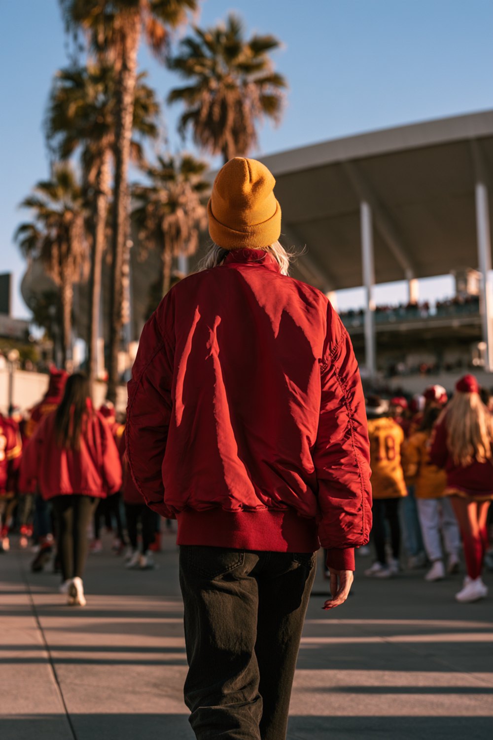 Fans in red and gold seen from behind walking toward a stadium past palm trees on a sunny day