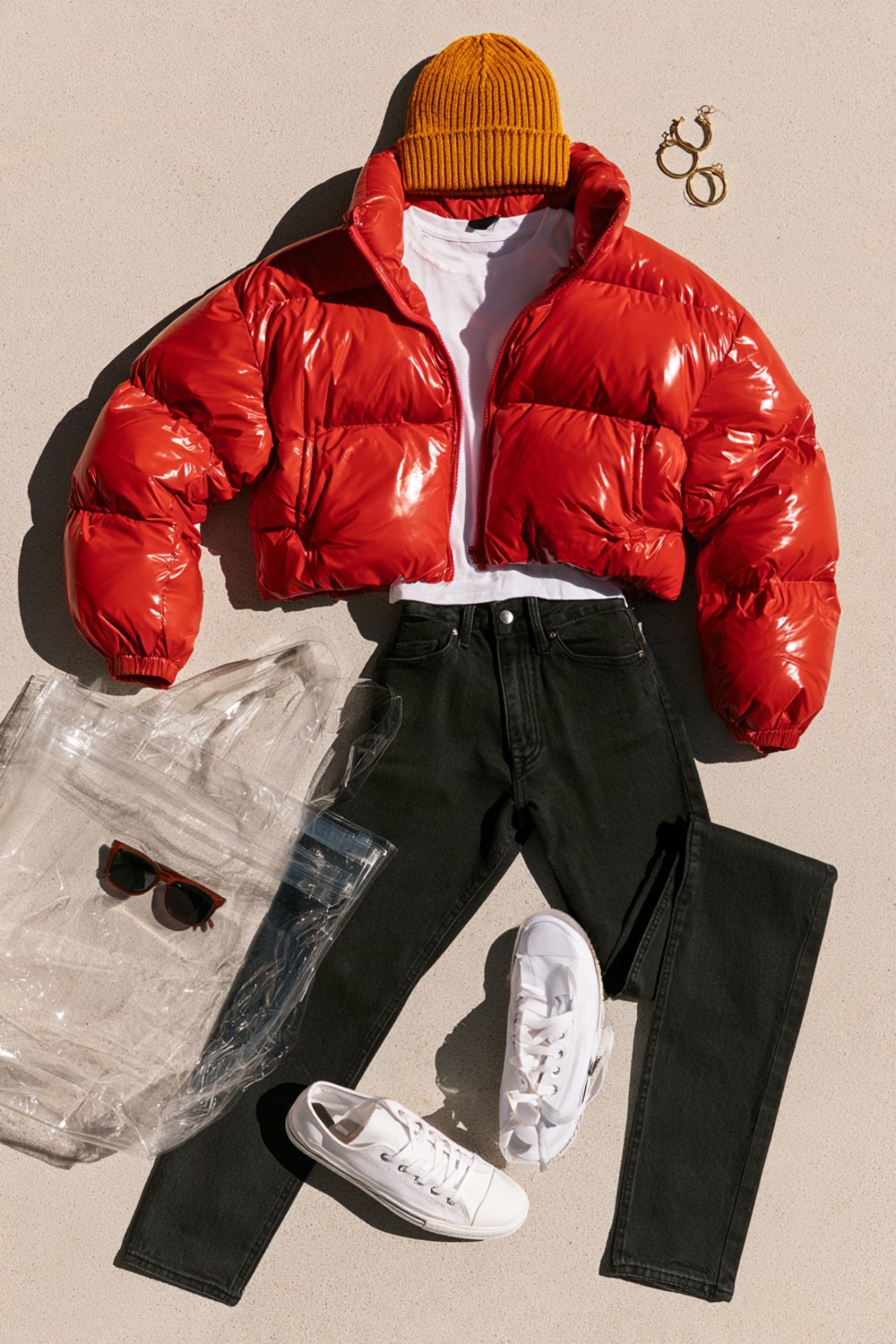 Another flat lay on concrete of a red puffer jacket, gold beanie, white tee, black jeans, white sneakers and a clear stadium tote