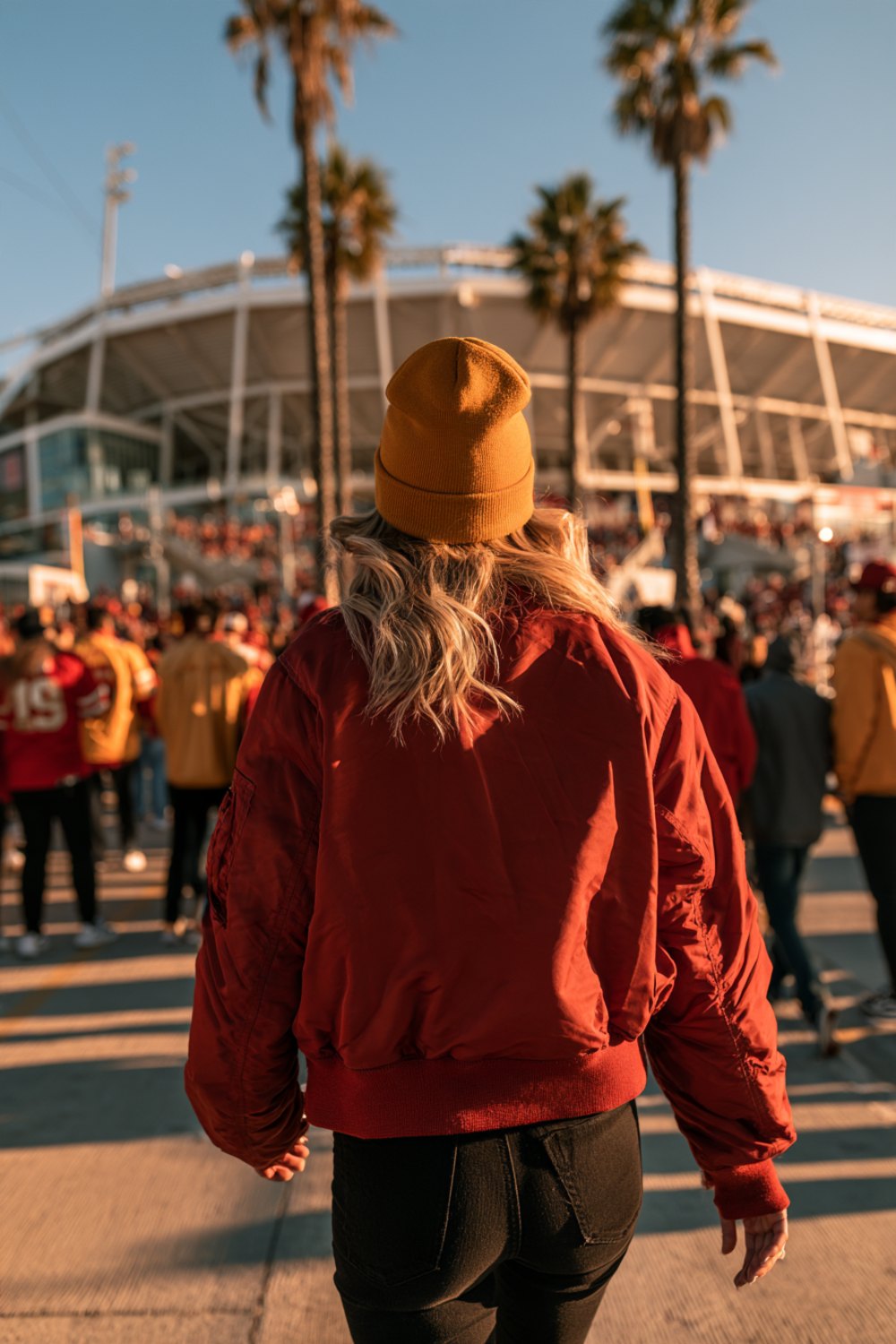 Fans in red and gold seen from behind walking toward a stadium past palm trees on a sunny day, another angle