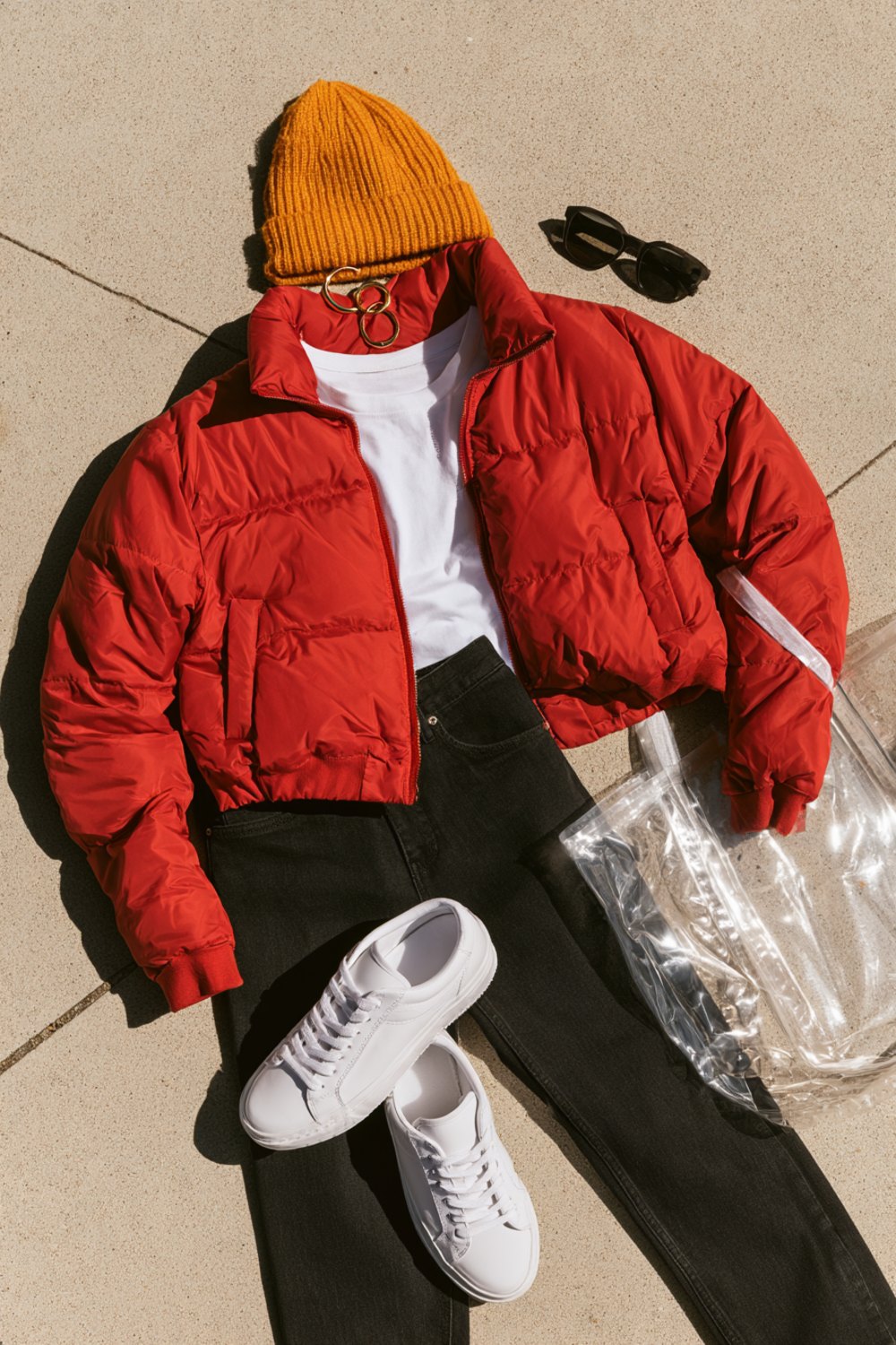 Another flat lay on concrete of a red puffer jacket, gold beanie, white tee, black jeans, white sneakers and a clear stadium tote