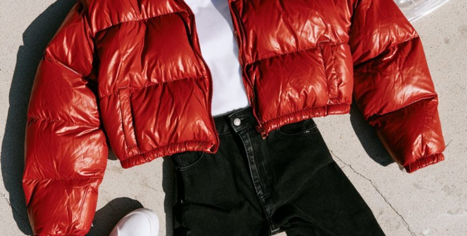 Flat lay on concrete of a red puffer jacket, gold beanie, white tee, black jeans, white sneakers and a clear stadium tote