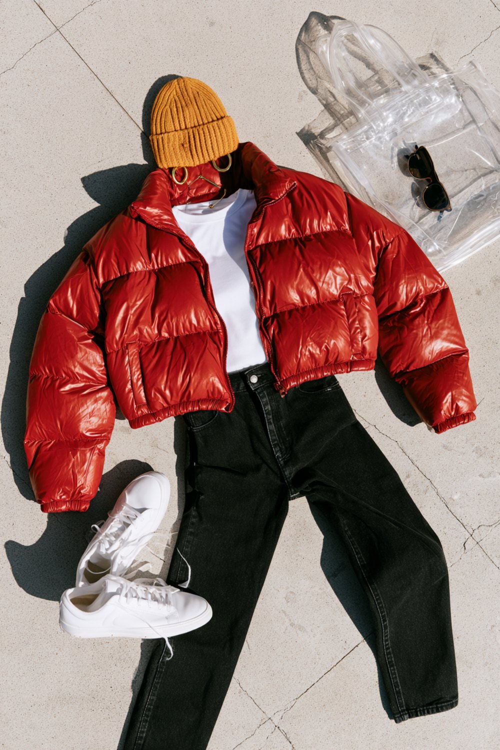Flat lay on concrete of a red puffer jacket, gold beanie, white tee, black jeans, white sneakers and a clear stadium tote
