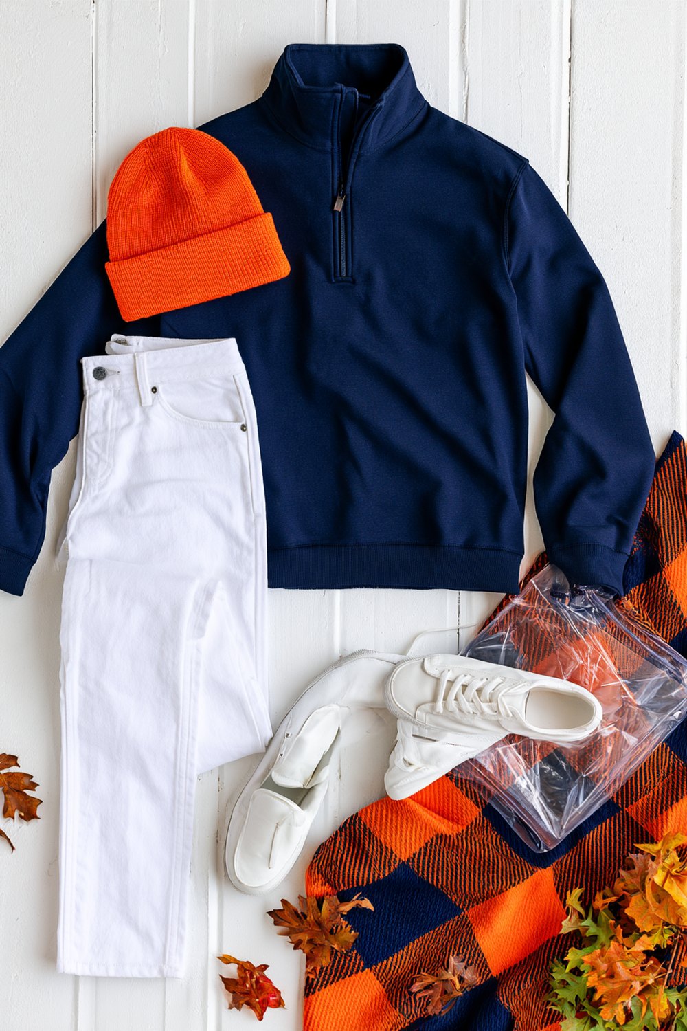 Auburn game day outfits flat lay with a navy quarter-zip, white jeans, orange beanie, white sneakers and a clear stadium bag