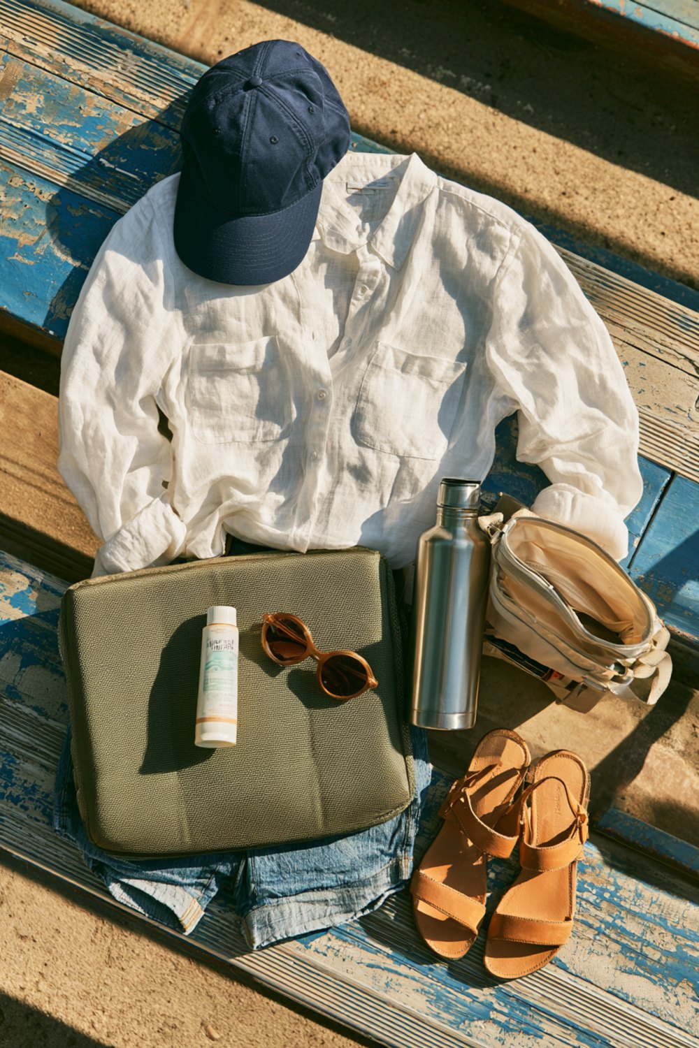 Flat lay on bleacher planks of a navy cap, white linen shirt, denim shorts, sandals, sunscreen and seat cushion