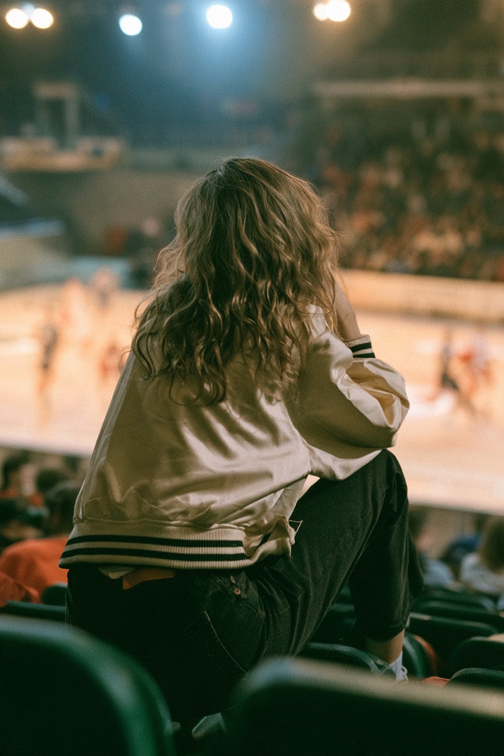 Woman seen from behind in an arena seat wearing an oversized cream varsity jacket and black jeans above a basketball court