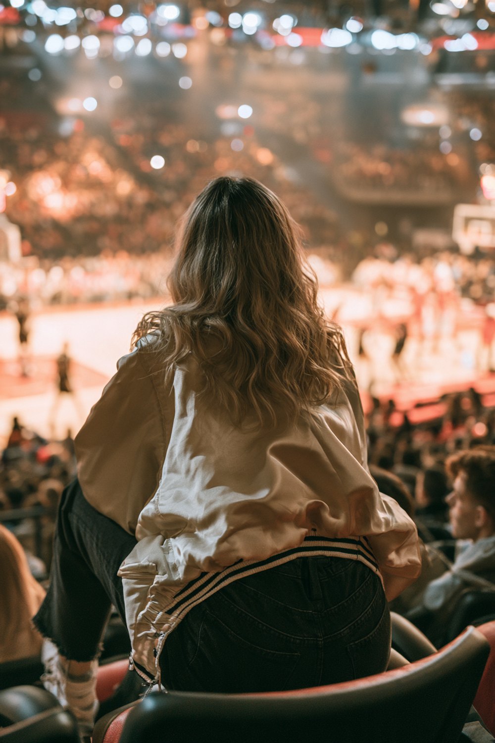 Fan from behind in a cream varsity jacket watching a basketball game from the upper seats