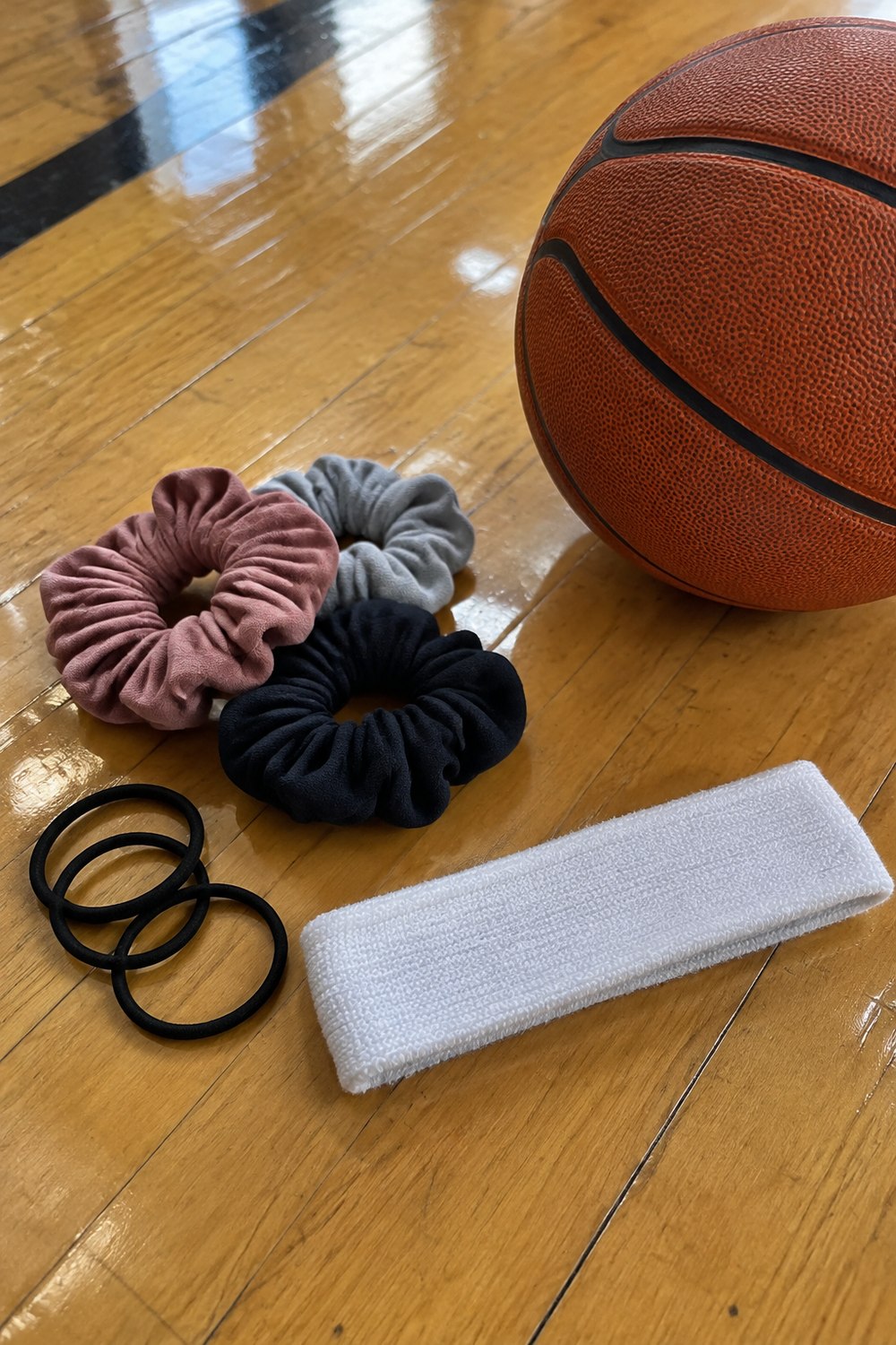 Soft scrunchies, black hair elastics and a plain white athletic headband next to a basketball on a gym floor