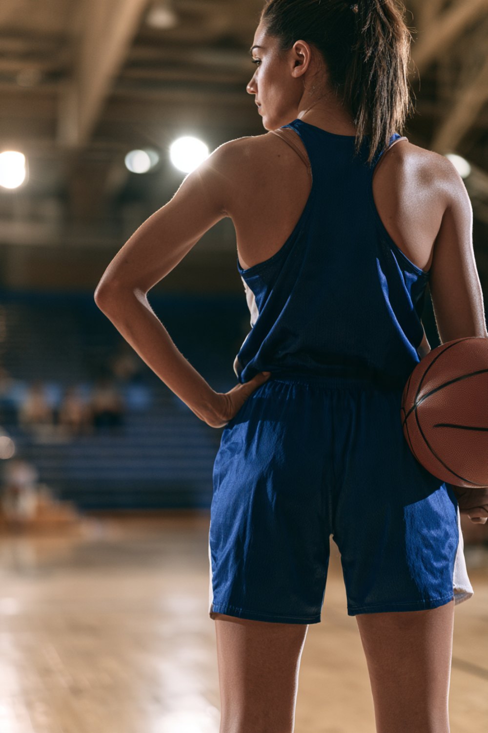 Basketball player seen from behind holding a ball on her hip in a blue uniform on a gym court