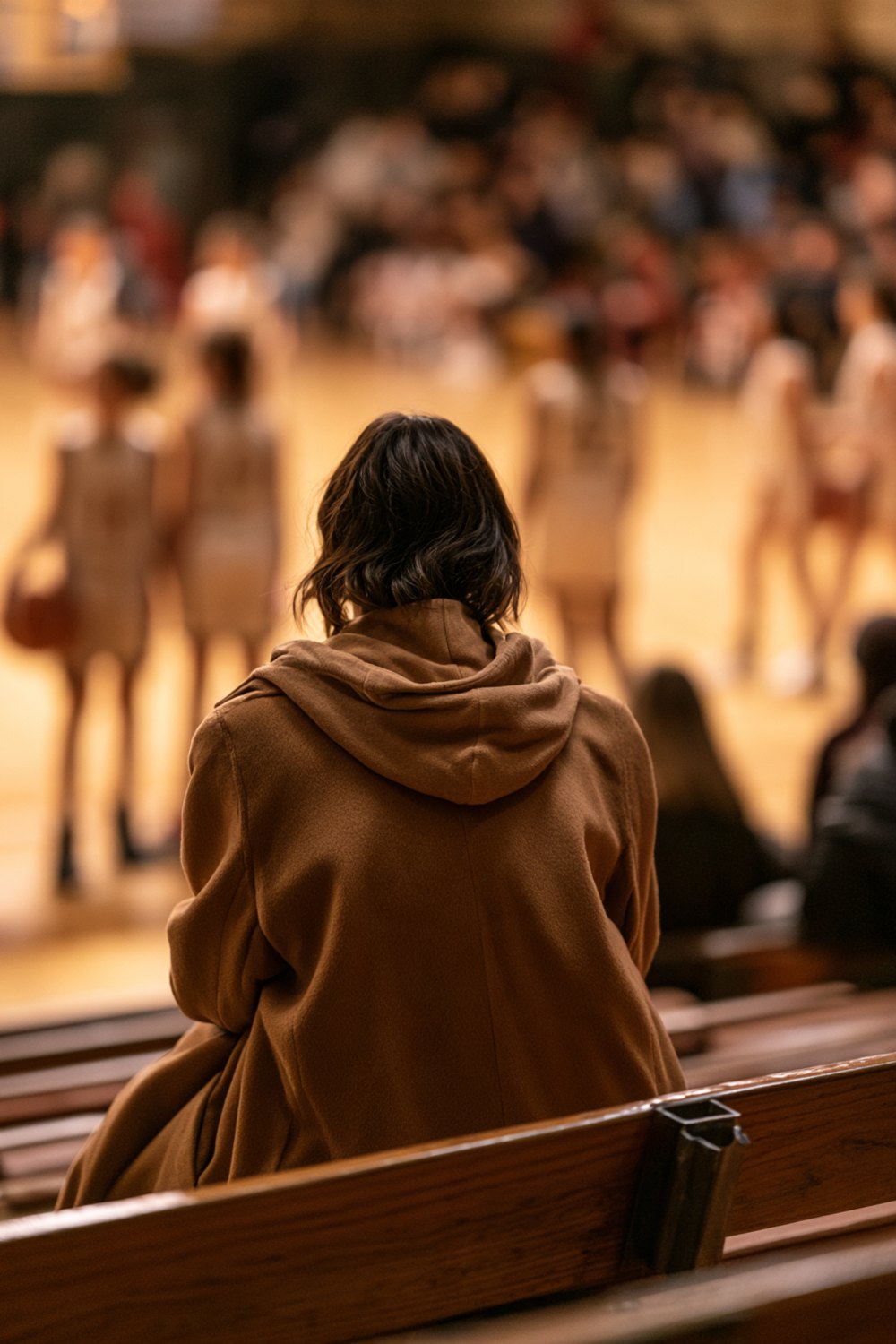 Woman in a camel hooded coat seen from behind on wooden gym bleachers during a youth basketball game