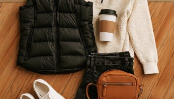 Flat lay on a hardwood court of a black puffer vest, cream half-zip, dark jeans, white sneakers and a tan crossbody bag