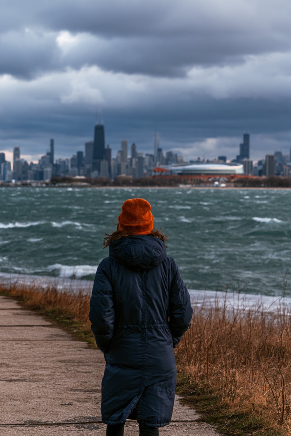 Person in a navy parka and orange beanie seen from behind on a windy lakefront path facing a city skyline, wider view