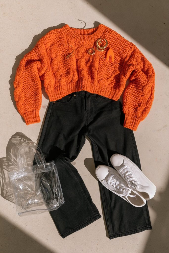 Flat lay of an orange chunky knit sweater, black wide-leg jeans, white sneakers and a clear stadium tote