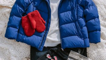 Flat lay of a royal blue puffer jacket, red knit beanie, red mittens, white thermal top and gray snow boots on a white knit blanket