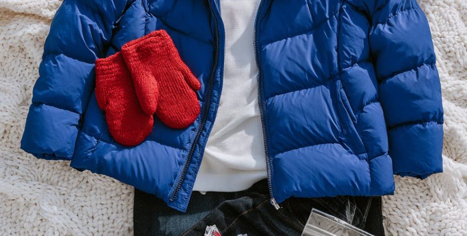 Flat lay of a royal blue puffer jacket, red knit beanie, red mittens, white thermal top and gray snow boots on a white knit blanket