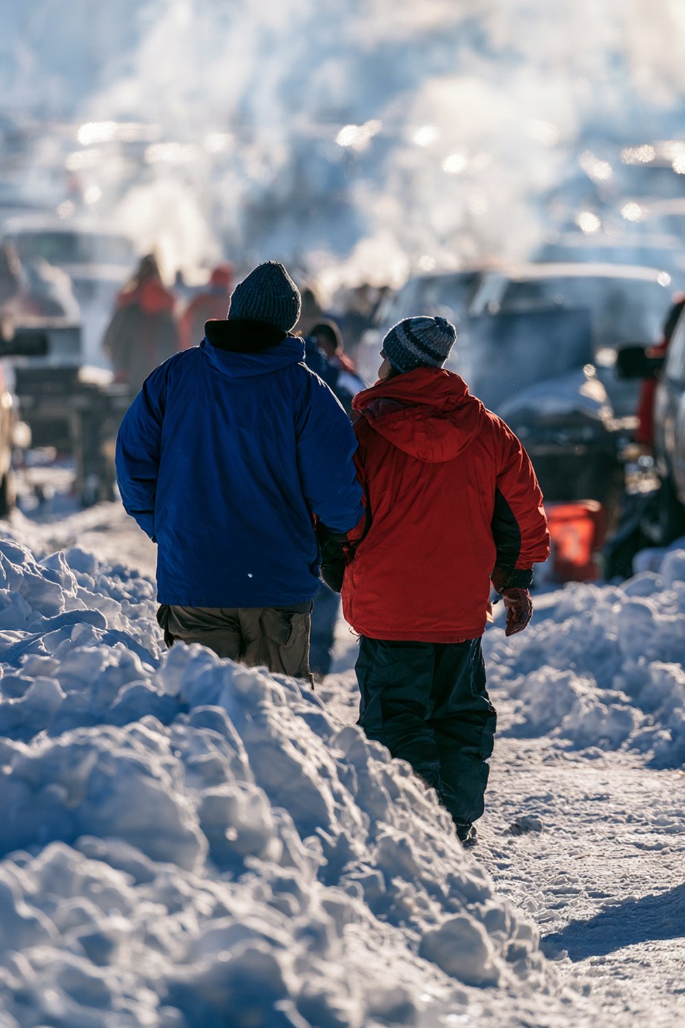 Two friends seen from behind walking through a snowy tailgate lot in royal blue and red winter jackets