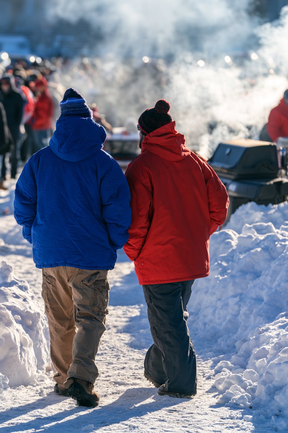 Two fans from behind in blue and red coats crossing a sunny, snowy parking lot past smoking grills