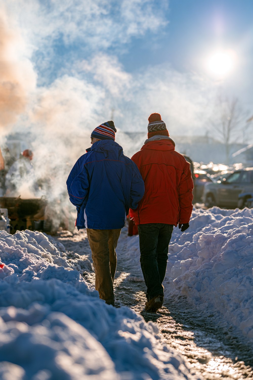 Back view of friends in blue and red jackets walking between snowbanks toward a tailgate