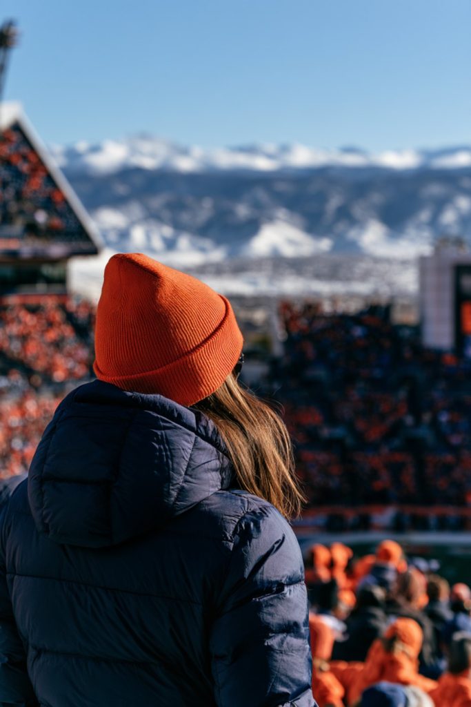 Fan in an orange beanie and navy puffer in the stands under a bright blue mile high sky