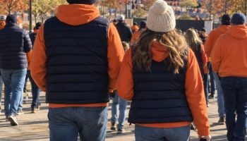 Couple in orange hoodies and navy vests walking to the stadium with mountains behind, seen from behind