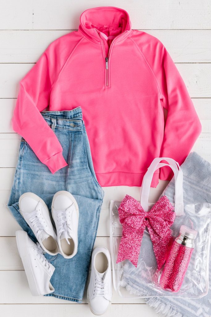 Pink pullover, jeans, white sneakers and a clear bag holding a sparkly bow laid out for a cheer weekend