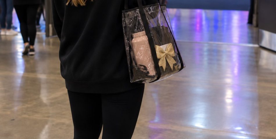 Cheer mom seen from behind in a black quarter-zip, leggings and white sneakers with a clear tote, walking toward a competition arena floor
