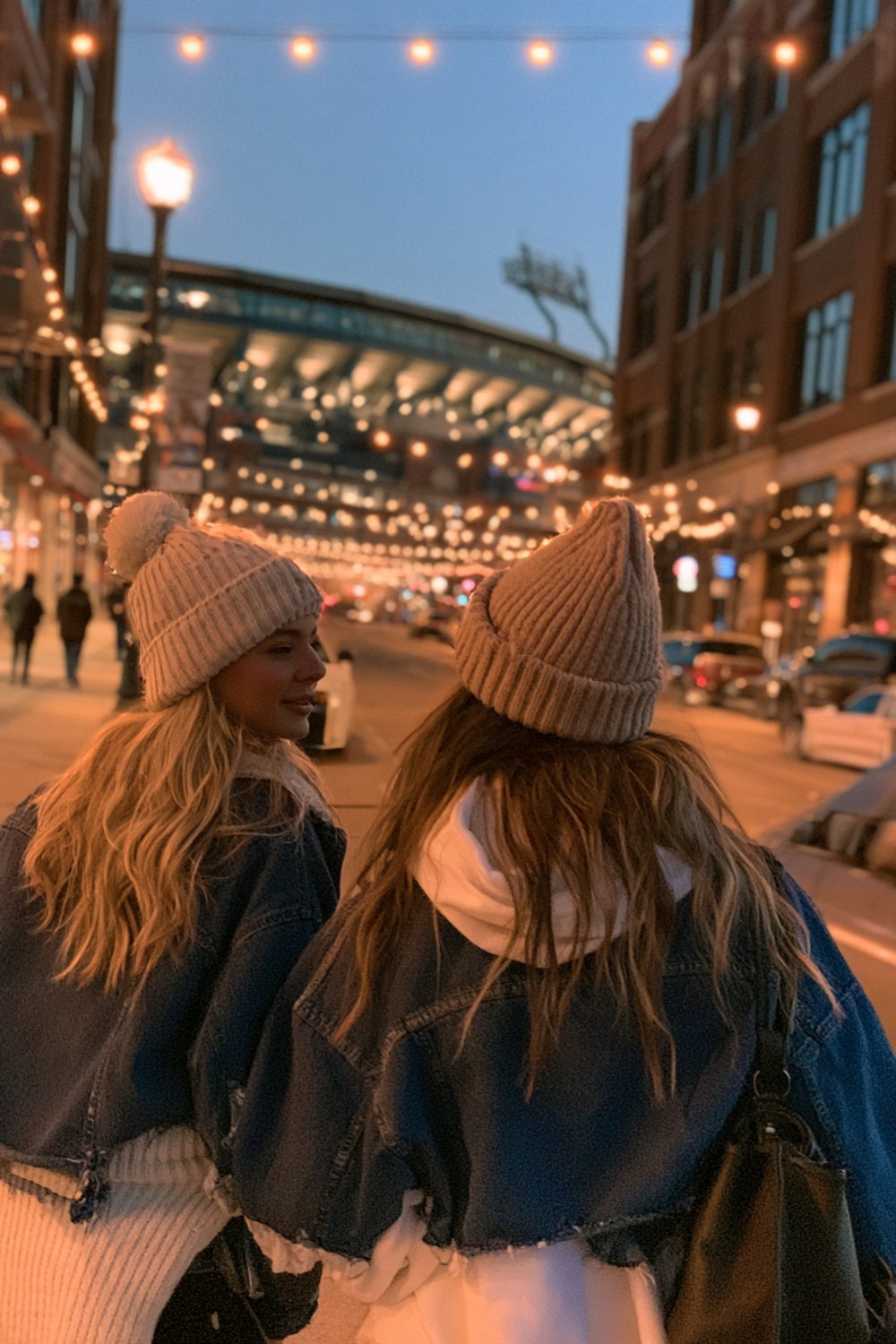 Two friends seen from behind in beanies walking down a string-lit downtown street toward a stadium at dusk