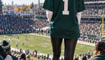 Woman in an open-air stadium wearing an oversized midnight green football jersey over a white long sleeve with black leather leggings and white sneakers, seen from behind