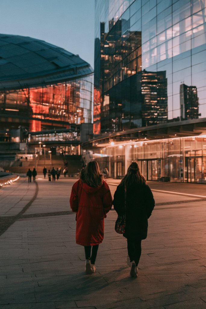 Two people in red and black walking past glass towers toward a downtown stadium
