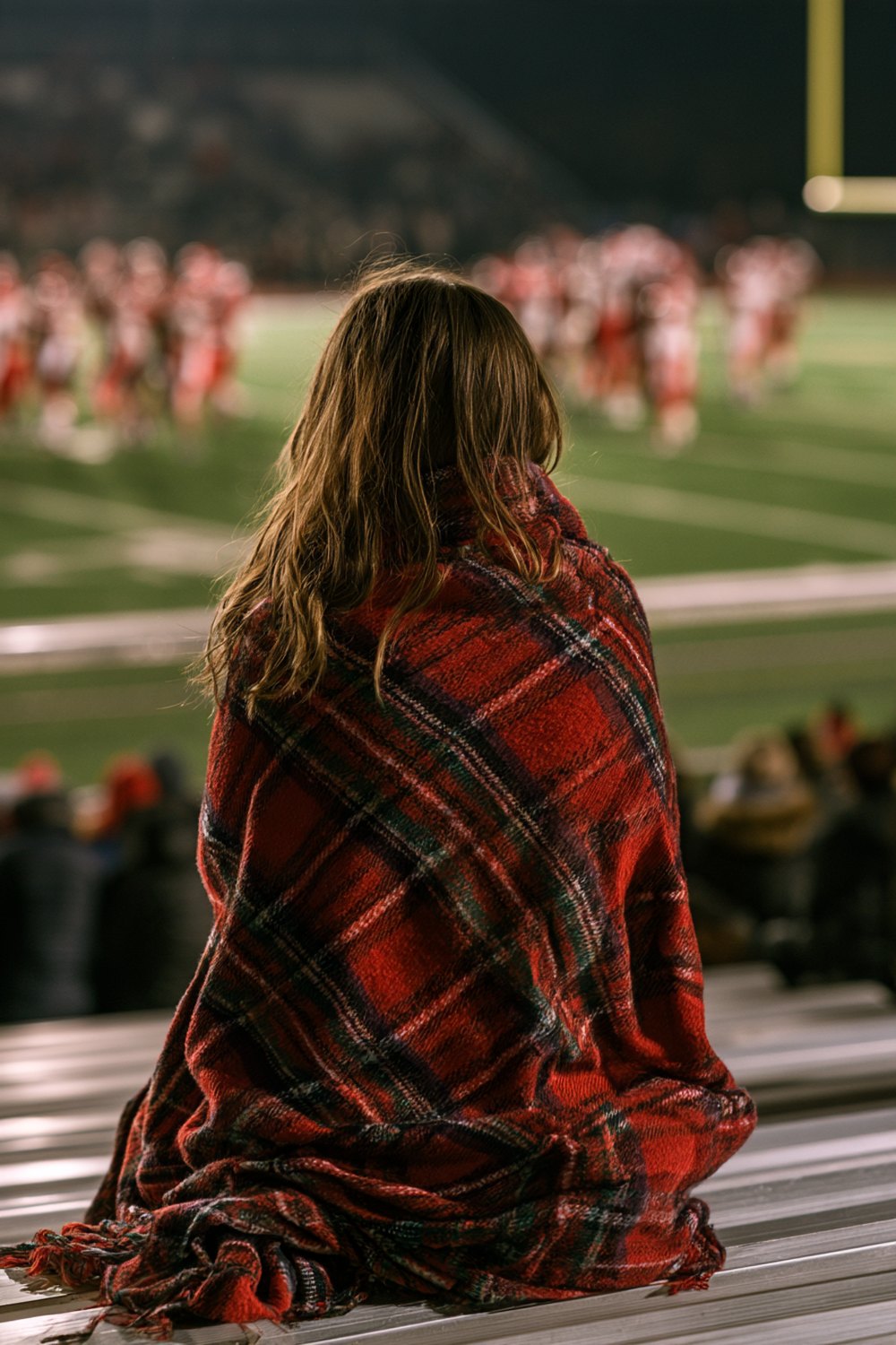 Football mom seen from behind on aluminum bleachers wrapped in a red plaid blanket under Friday night lights