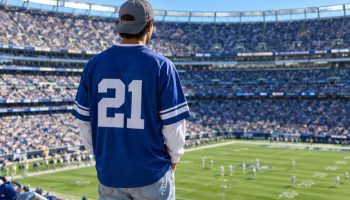 Game day outfits for men: a man seen from behind in a stadium wearing an oversized royal blue number 21 jersey over a white long sleeve, light baggy jeans, white sneakers and a backwards grey cap