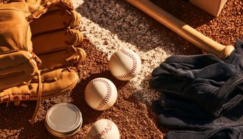 Flat lay on infield dirt of a leather baseball glove, baseballs, wooden bat, batting gloves and a wrapped gift