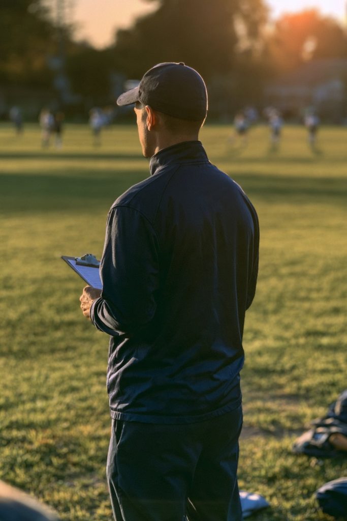 Coach seen from behind on a sunny sideline holding a clipboard