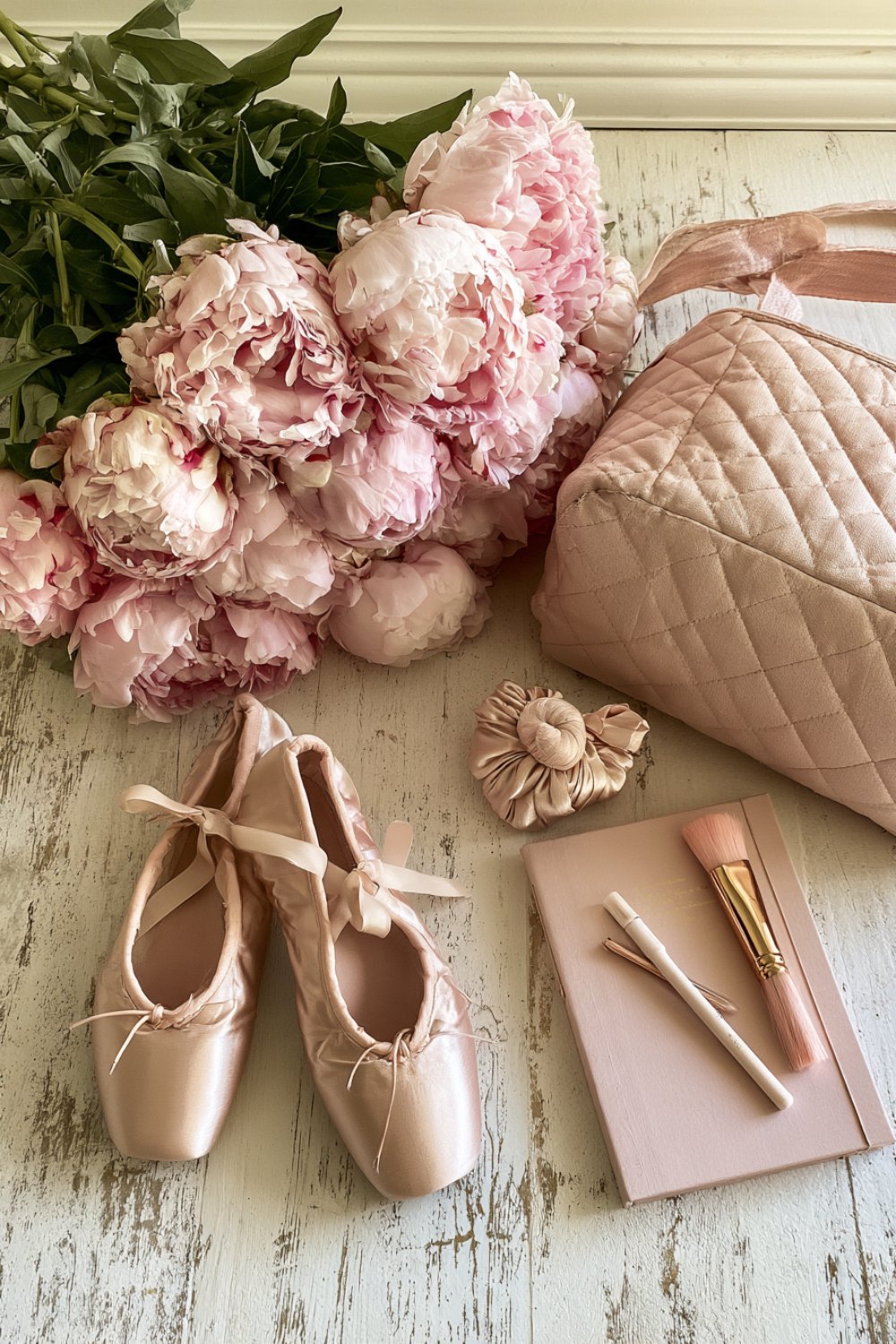 Gifts for a dancer laid out on pale wood: ballet slippers, pink peonies, a quilted bag and a journal