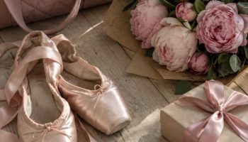 Flat lay of pink satin ballet slippers, a peony bouquet in kraft paper, a quilted dance bag, bobby pins and a gift box with a satin bow