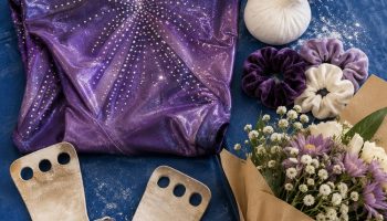 Flat lay on a blue gymnastics mat with a sparkly purple leotard, leather grips, a chalk ball, scrunchies, a water bottle and a small bouquet