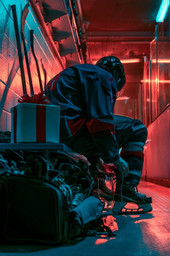 Hockey player seen from behind lacing skates in a locker room next to a gift box