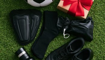 Flat lay of soccer gift ideas on green turf: a black and white soccer ball, shin guards, socks, black cleats, a water bottle and a gift box with a red bow