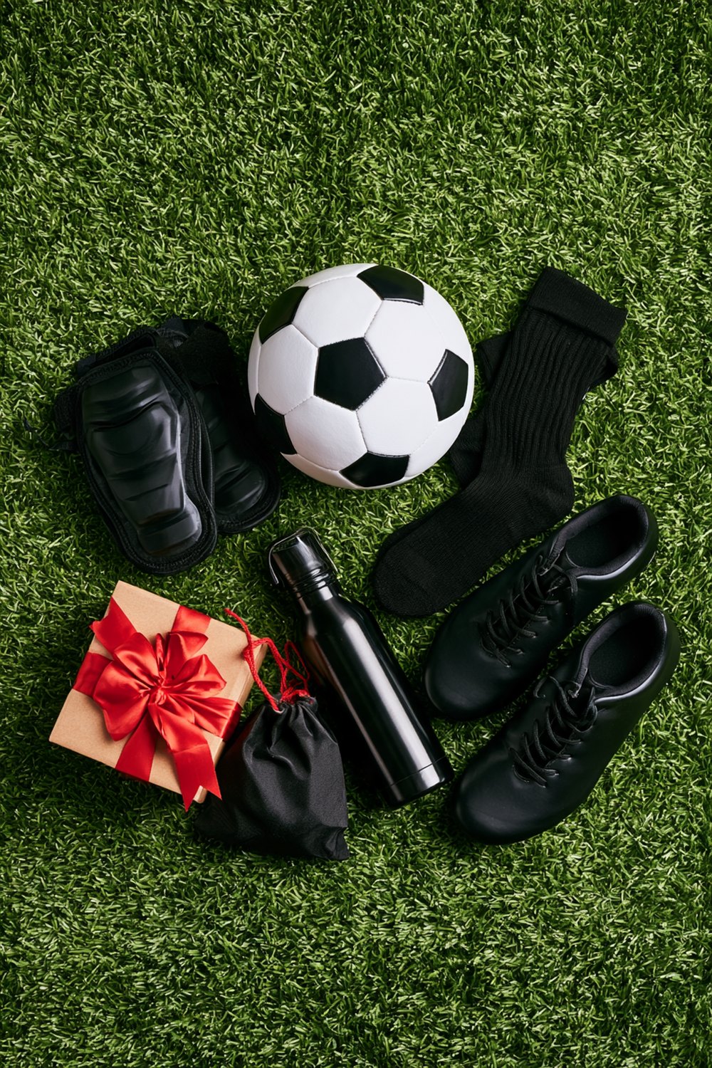 Soccer gifts on artificial turf including shin guards, a ball, black socks, plain black cleats, a drawstring bag and a wrapped present