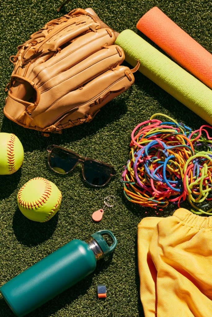 Flat lay on turf of a leather fielding glove, yellow softballs, bat grip, sunglasses and colorful hair ties