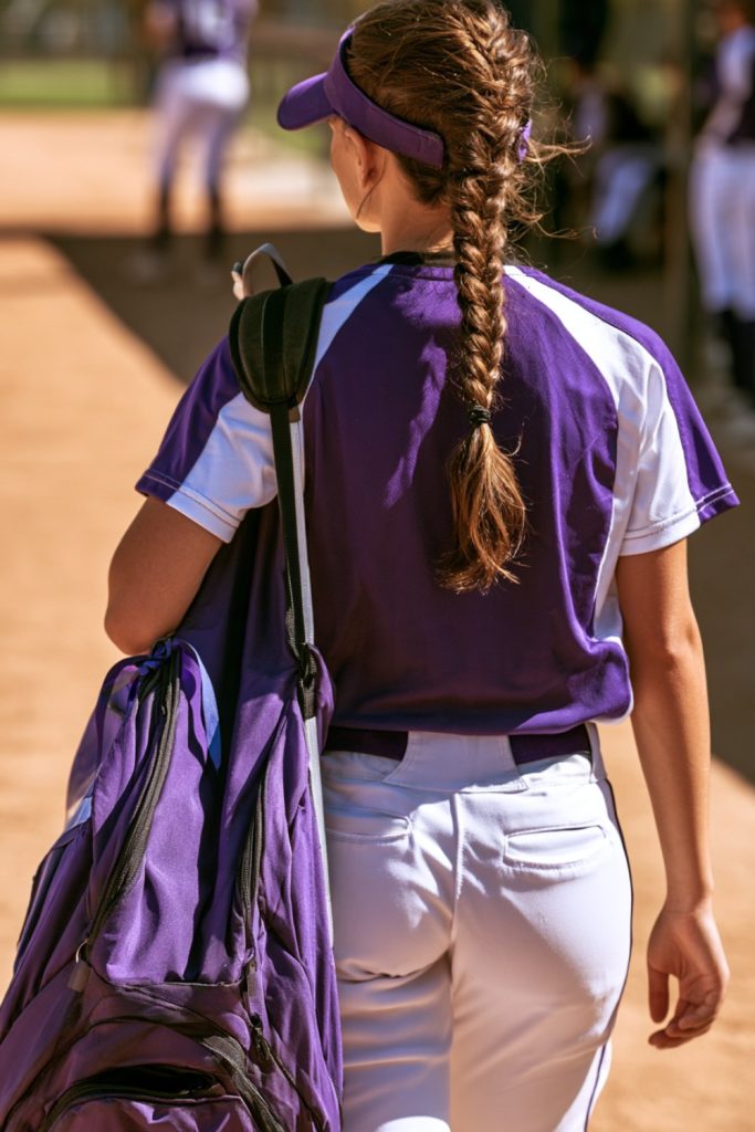 Softball player in a purple uniform seen from behind carrying her bat bag toward the field