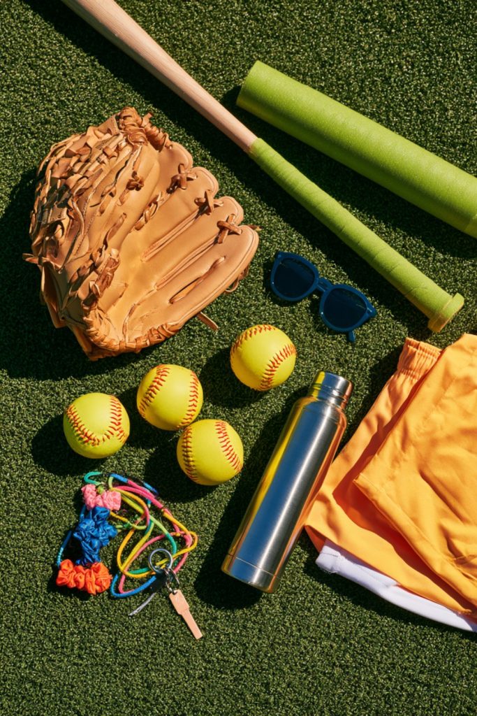Softball glove, bat, softballs, water bottle and sliding shorts laid out on green turf as gift ideas