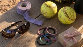 Dugout bench with a worn softball glove, two yellow fastpitch softballs, grip tape, sunglasses, hair ties and a small gift box
