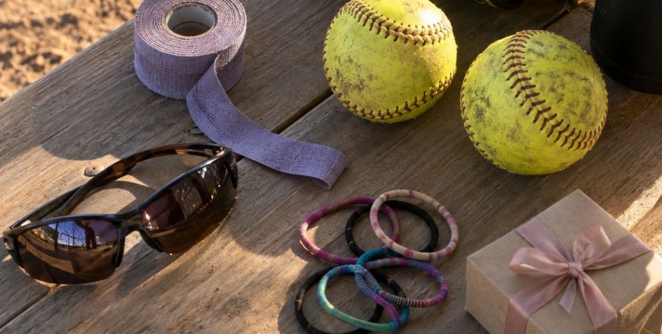 Dugout bench with a worn softball glove, two yellow fastpitch softballs, grip tape, sunglasses, hair ties and a small gift box