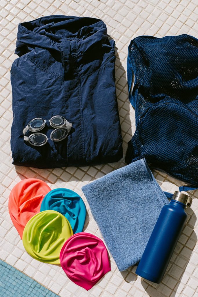 Flat lay of swimmer gifts: navy swim parka, goggles, colorful silicone caps, towel and water bottle on pool tiles